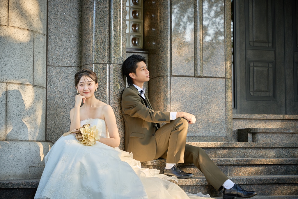 Bride in a white ball gown holding a bouquet rests beside her groom in a golden-brown suit on the stone steps of a grand historic building.
