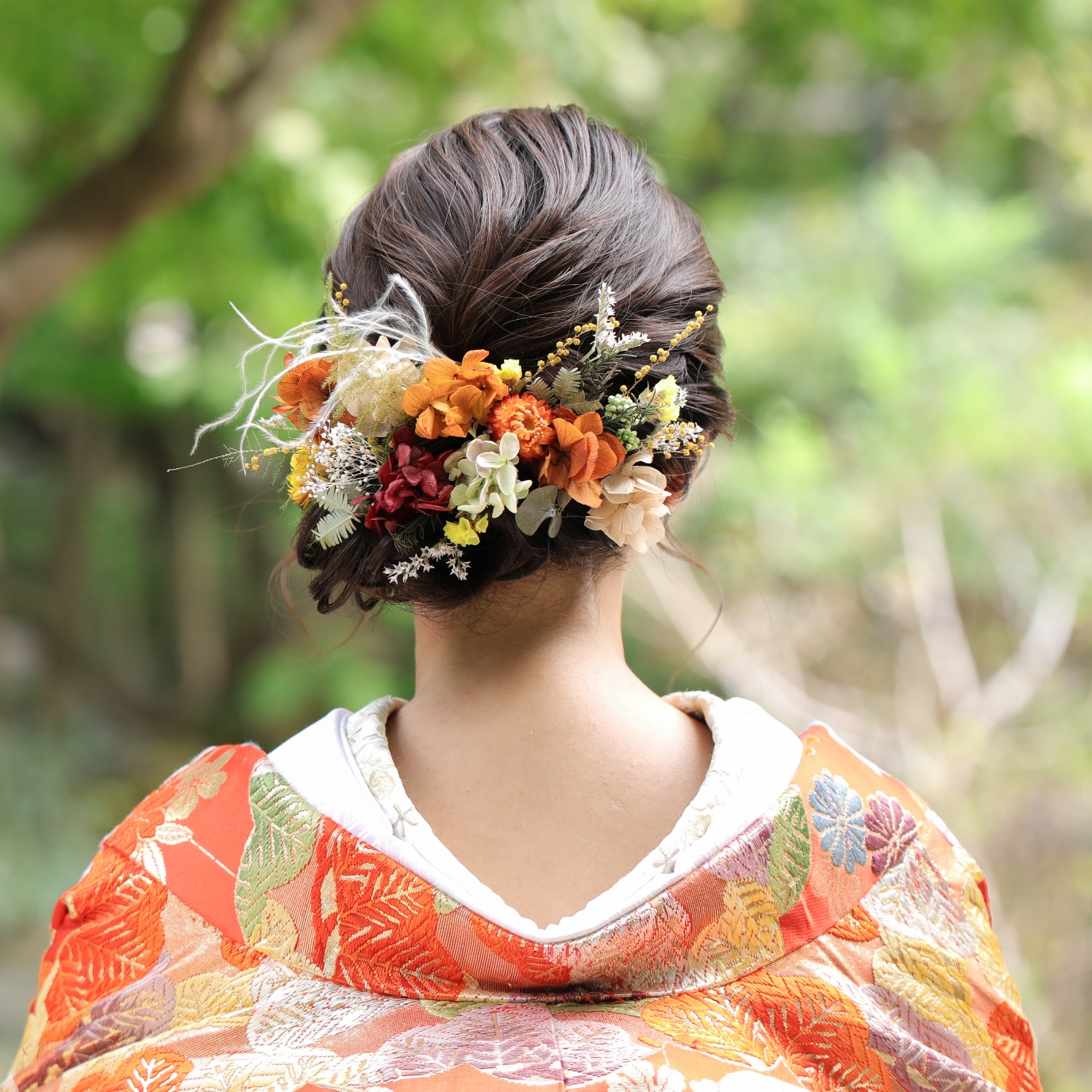 A back view of a bridal updo woven with vivid orange, red, and cream dried flowers, worn with a richly embroidered orange iro-uchikake kimono.