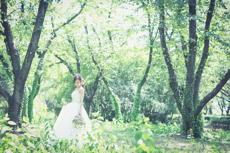 Bride standing amid a lush green grove of trees at Shonai Ryokuchi Park, holding a small bouquet.