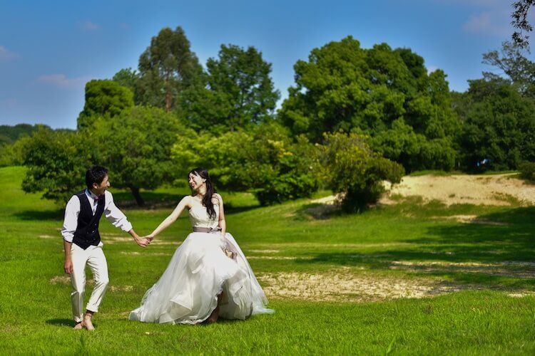 Playfully leading each other across the open lawns of Odaka Ryokuchi Park, a favorite Nagoya wedding photography spot framed by lush green trees under a clear blue sky.