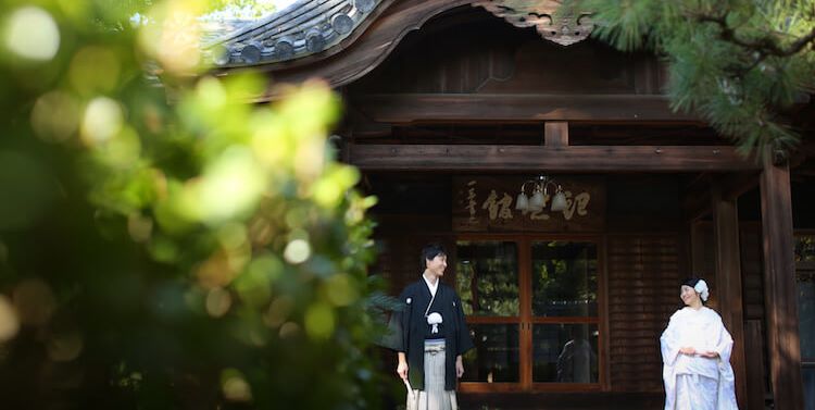 Couple in kimono standing before a traditional building with an ornate carved gable at Nakamura Park's Toin Chaseki hall.