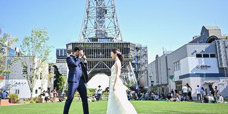 Couple facing each other on a green lawn beneath the towering Nagoya TV Tower at Hisaya Odori Park.