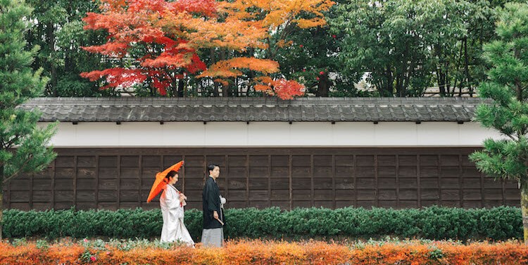 Couple in kimono walking past a long wooden fence beneath vivid red and orange autumn maples at Tokugawaen Garden.
