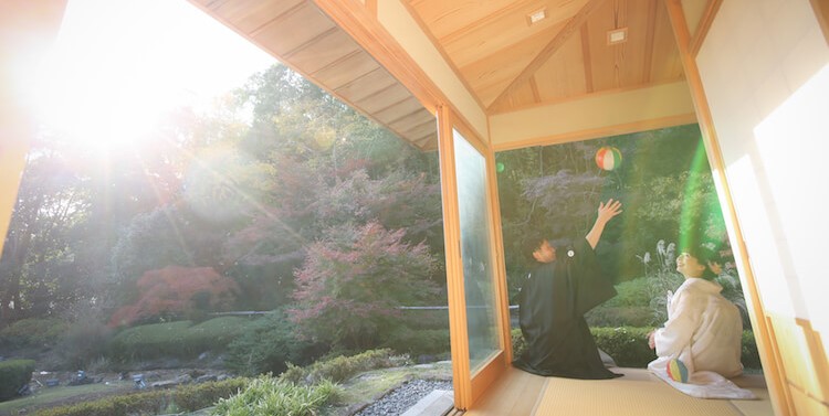 Couple in kimono kneeling in a sunlit wooden pavilion tossing a paper ball, autumn maples glowing outside at Sosetsu-an.