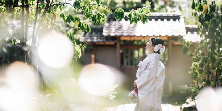The bride gazes up thoughtfully in the garden at Kakutei teahouse, traditional tiled roofs visible through the trees.