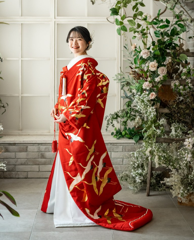 Bride in a red uchikake with gold and white cranes, standing by an arched window framed with fresh flowers at our Yokohama studio — kimono wedding photos in Japan (Tsuruki / YB13)