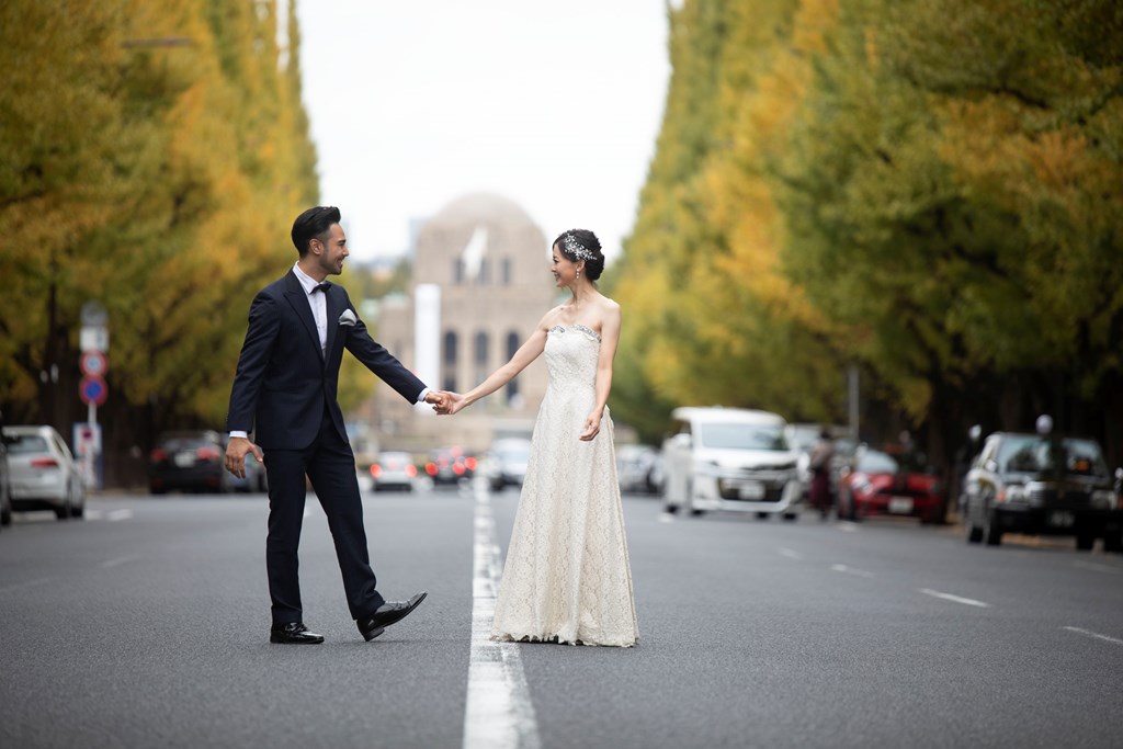 Bride in a white wedding dress and groom in a navy tuxedo walking hand in hand down the center of the Jingu Gaien Ginkgo Avenue