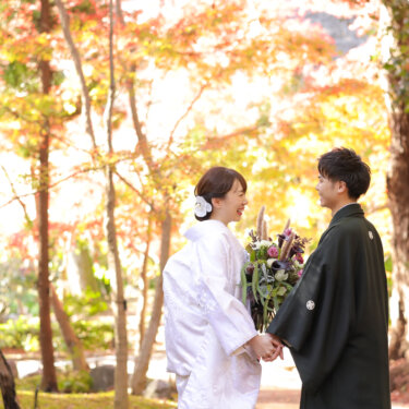 Bride in shiromuku holding a bouquet and groom in montsuki hand in hand under golden autumn trees in a Japanese garden, a fall foliage kimono wedding photography