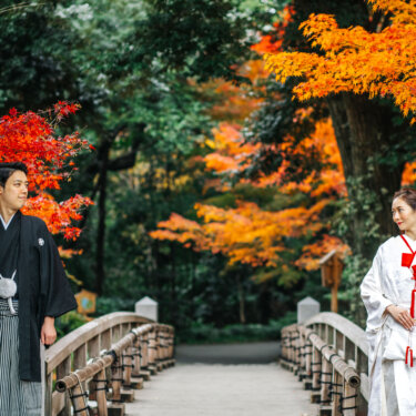 Groom in montsuki hakama and bride in white shiromuku facing each other across a wooden bridge framed by red and orange maple leaves, an autumn kimono pre-wedding photoshoot