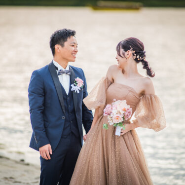 Groom in a navy tuxedo and bride in a blush gown with a pink bouquet smiling at each other on the sandy shore, Japan wedding photography