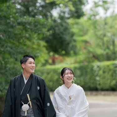 Bride in shiromuku and groom in montsuki and hakama walking and laughing along a green path in Daisen Park, a natural walking shot from their pre-wedding photos in Osaka