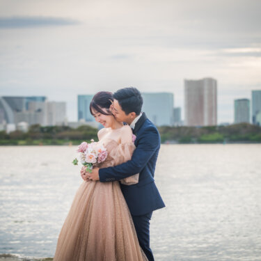 Groom hugging the bride from behind and kissing her cheek with the Tokyo waterfront skyline in the background, Odaiba wedding photos