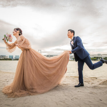 Groom playfully lifting the long train of the bride's blush gown on the beach, a fun pre-wedding photoshoot pose
