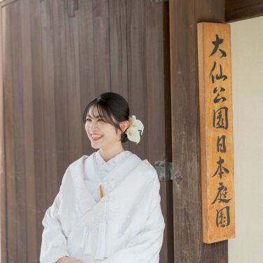 Smiling bride in shiromuku with a white orchid in her hair standing by the wooden gate sign of Daisen Park Japanese Garden, a bridal portrait from a kimono wedding photoshoot in Sakai