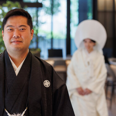 Groom in black montsuki smiles at the camera with the bride in shiromuku and wataboshi softly blurred behind him, Japan wedding photography
