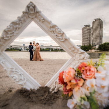 Couple framed by a white ornate frame on the sand with the Rainbow Bridge behind them, a creative prop idea for Odaiba pre-wedding photography
