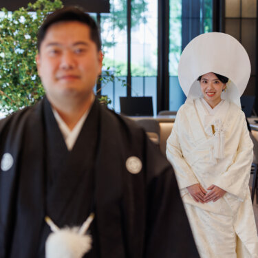Groom in montsuki stands blurred in the foreground while the bride in a round wataboshi hood smiles behind him, a creative Japanese wedding photoshoot