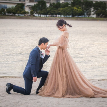 Groom kneeling on the beach to kiss the bride's hand as her long train spreads out, a romantic proposal pose for a Japan wedding photoshoot