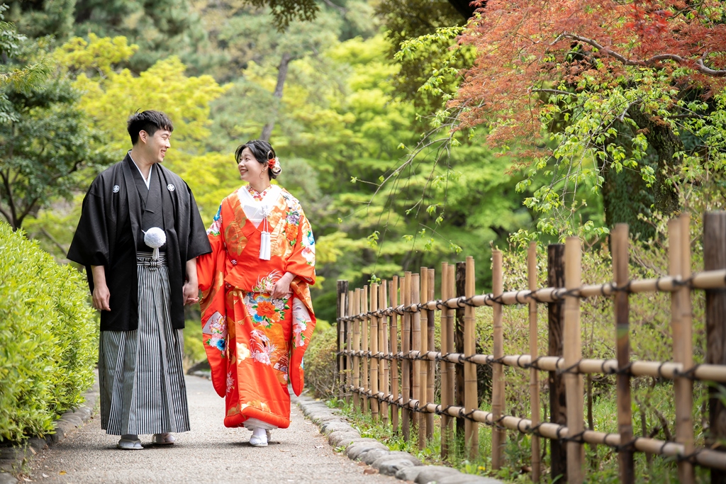 Bride in a red iro-uchikake and groom in black montsuki hakama walking along the bamboo fence path at Kansenen Garden, Tokyo kimono wedding photos