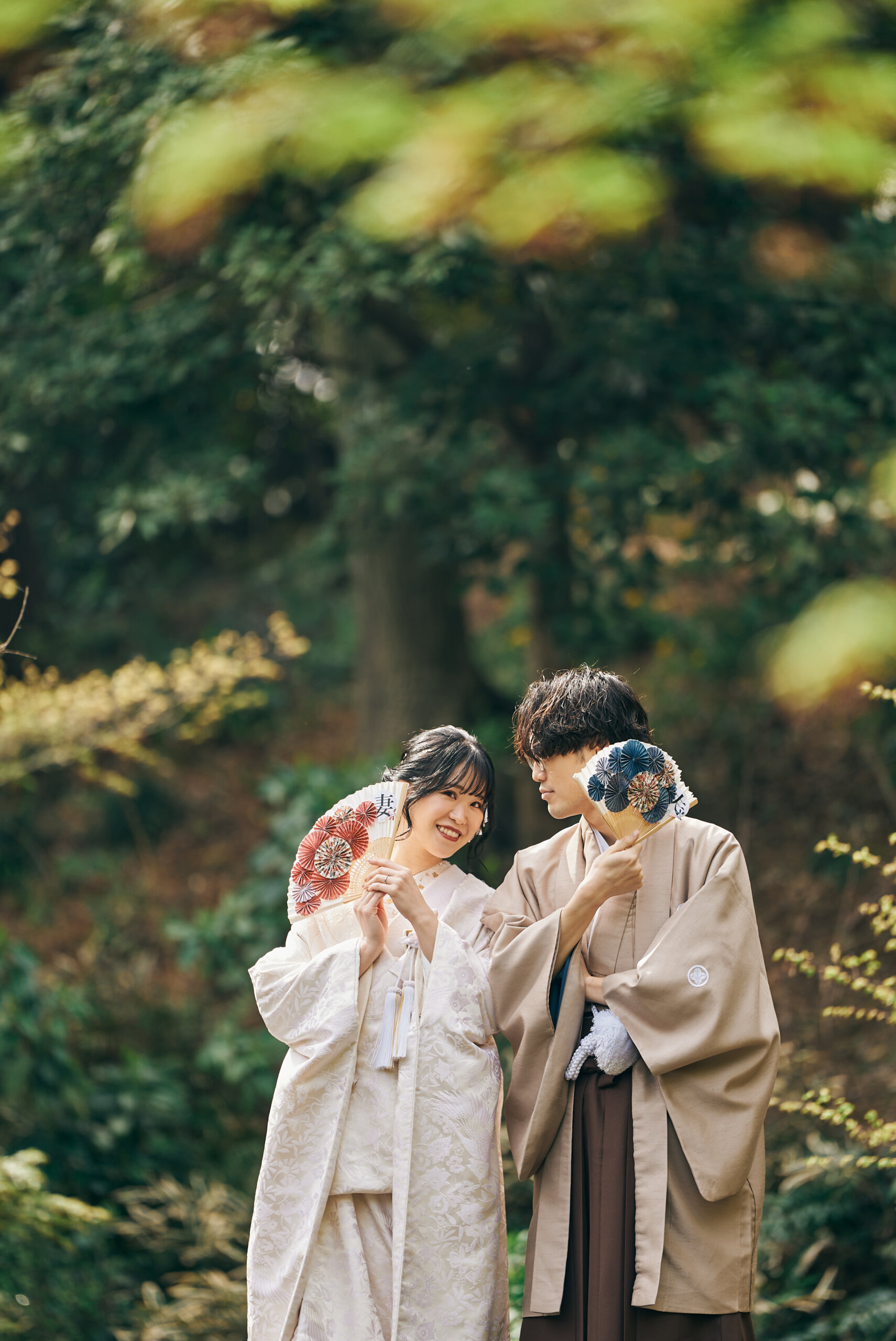 Bride in shiromuku and groom in kimono covering their faces with colorful folding fans at Kansenen Garden, a playful kimono wedding photo pose