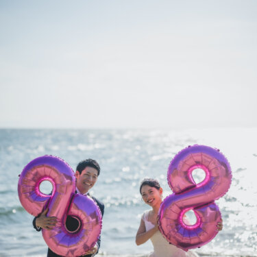 Laughing groom in a tuxedo and bride in a wedding dress each holding pink number balloons by the sea in Chita Peninsula wedding photos