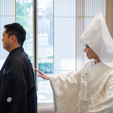 Bride in shiromuku and a pointed wataboshi hood reaches out to tap the groom's shoulder from behind, a first look style kimono wedding photo
