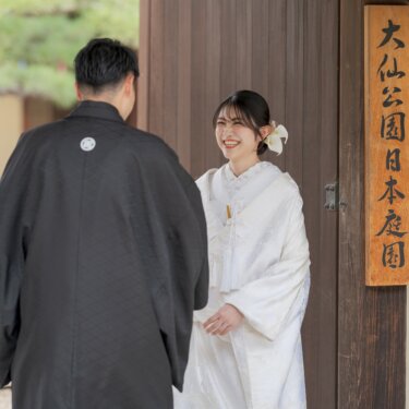 Bride in shiromuku smiling at the groom in black montsuki, seen from behind, at the entrance gate of Daisen Park Japanese Garden in Sakai, Osaka, a candid wedding photography moment