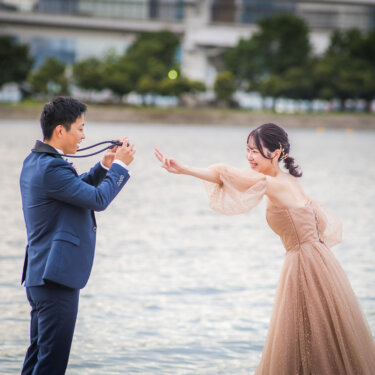 Groom taking a photo of his bride with a film camera as she strikes a playful pose by the water, a fun pre-wedding photoshoot idea