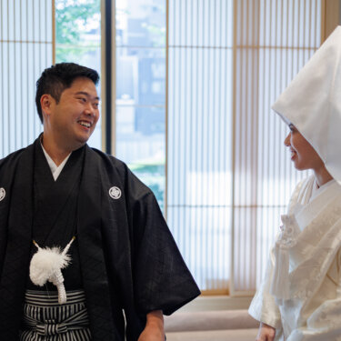 Groom in black montsuki hakama laughs while the bride in shiromuku and a pointed wataboshi hood looks at him, natural kimono wedding photography