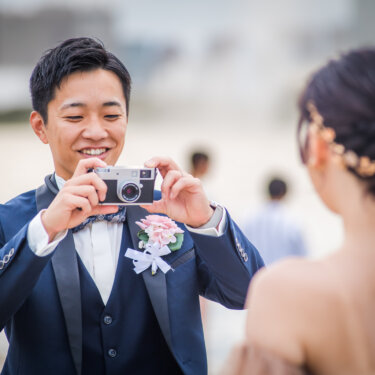 Groom in a navy tuxedo snapping a picture of his bride with a vintage camera, seen over her braided updo, in Odaiba wedding photos