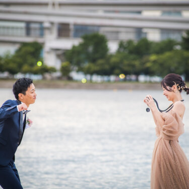 Bride and groom pointing cameras at each other across the shoreline at Odaiba Seaside Park, a playful prop idea for wedding photos in Tokyo