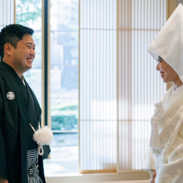 Groom in dark montsuki laughs as the bride in a pointed wataboshi hood turns toward him by the lattice screens, a candid Japanese wedding photoshoot