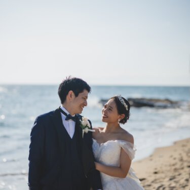 Groom in a navy tuxedo and bride in an off-shoulder wedding dress smiling at each other on the sand, with a bouquet in hand for their beach wedding photos