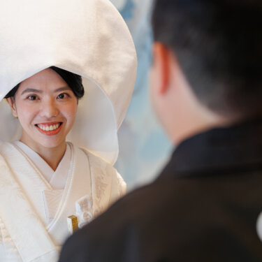Over-the-shoulder shot of the groom in montsuki facing the smiling bride in a round wataboshi hood, a sweet kimono wedding photos moment