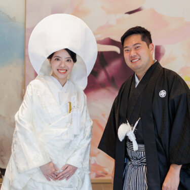 Bride in a round wataboshi hood and groom in montsuki hakama smile in front of a pastel floral mural, a cheerful Japanese studio wedding photoshoot