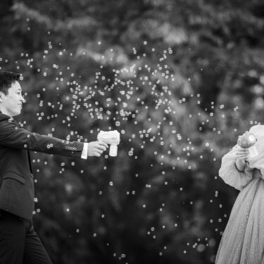 Black and white photo of the groom and bride playfully spraying bubble guns at each other among the trees, a creative idea for wedding photos in Japan