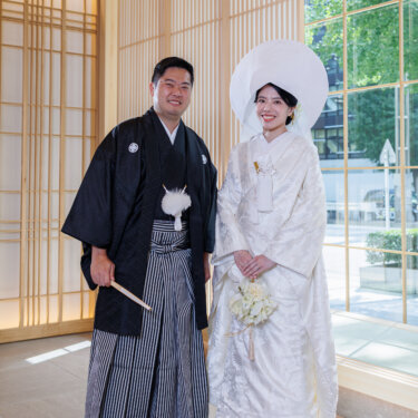 Groom in montsuki hakama and bride in shiromuku and wataboshi with a white bouquet stand together near a sunny window, Japan wedding photography