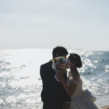 Bride and groom hiding a kiss behind the bouquet against the glittering sea, a romantic pose idea for pre-wedding photography in Aichi
