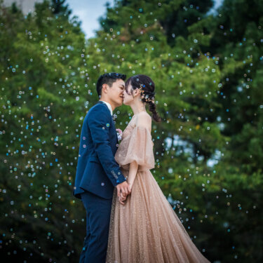 Couple kissing among floating bubbles in front of lush green trees, a dreamy moment from their pre-wedding photoshoot