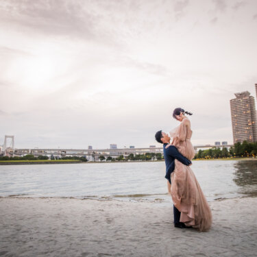 Groom lifting the bride high on the sand, with the Rainbow Bridge and Odaiba skyline behind them, a romantic pose for Tokyo pre-wedding photography