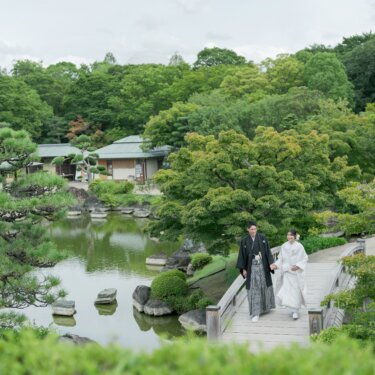 Wide view of the bride and groom in kimono walking over a footbridge by the pond, surrounded by pines and the traditional pavilions of Daisen Park Japanese Garden, a scenic pre-wedding photo