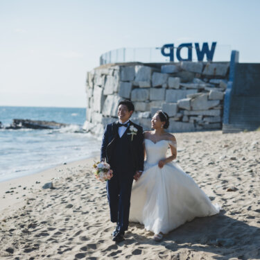 Couple walking hand in hand along the sandy beach beside a stone seawall near Noma Lighthouse during their wedding photoshoot