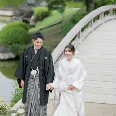 Bride in shiromuku and groom in montsuki and hakama holding hands and smiling as they walk from a wooden arched bridge by the pond, a natural kimono wedding photography shot