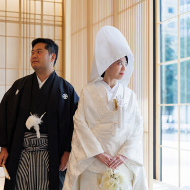 Groom in montsuki hakama and bride in shiromuku with a pointed wataboshi hood stand side by side looking away, an elegant kimono wedding photo