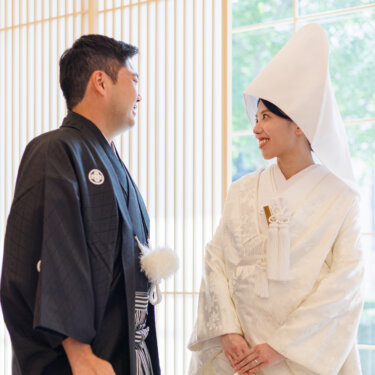 Groom in montsuki hakama and bride in a pointed wataboshi hood smile at each other by tall lattice screens, Japanese wedding photography