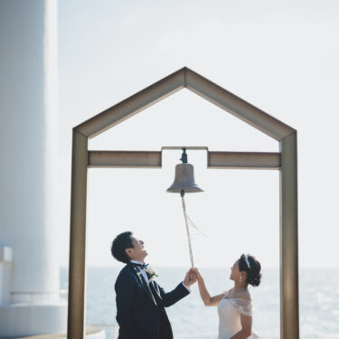Bride and groom ringing the bell under the house-shaped frame next to Noma Lighthouse, a popular spot for wedding photography on the Chita Peninsula