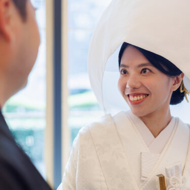 Bride in a white wataboshi hood beams at the groom in the foreground, a candid moment from a kimono wedding photoshoot in Japan