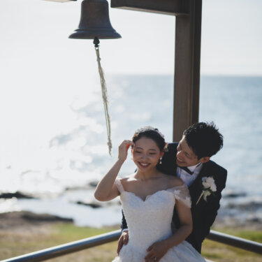 Groom hugging the bride from behind as she smiles and touches her hair beneath the lighthouse bell, a natural pose for Japan wedding photos