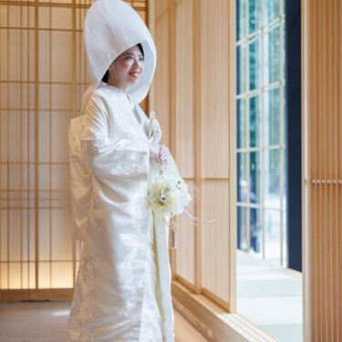 Bride in shiromuku and a round wataboshi hood holds a white bouquet and smiles by the window, a full-length Japan wedding photography portrait