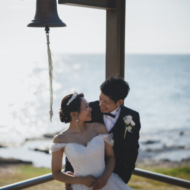 Smiling couple in a back hug under the bell by the sea at Noma Lighthouse, the bride in an off-shoulder lace wedding dress and tiara in their wedding photos